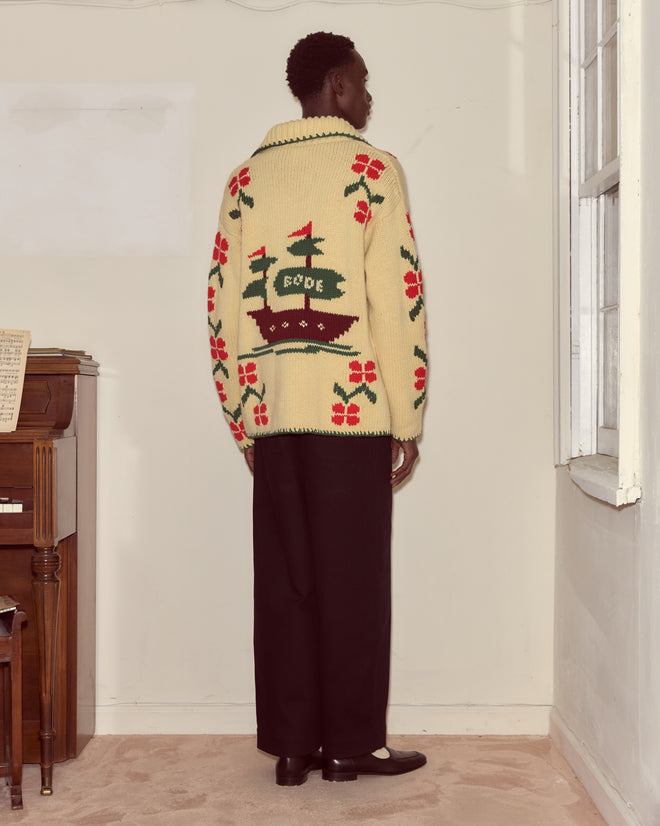 A person is standing in a room near a piano and window, wearing the cream Pirate Ship Cardigan, which features a ship and “BODE” on the back.