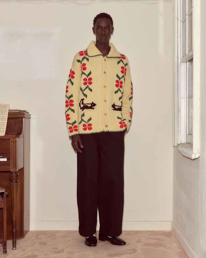 Wearing the Pirate Ship Cardigan with black pants, a person stands in a minimal room next to a piano and window.