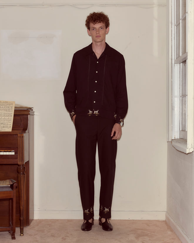 A man with curly hair wears the Pointer Long Sleeve Shirt, which is black with ladderwork detailing, and stands beside a piano in a softly lit room.