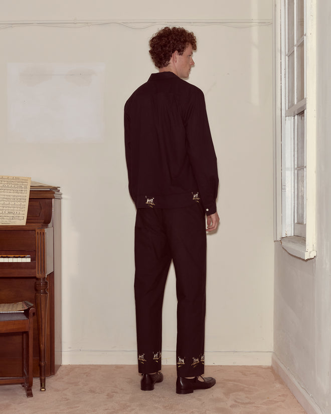 A man wearing the Pointer Long Sleeve Shirt stands facing a corner near a window and an upright piano with sheet music on top.