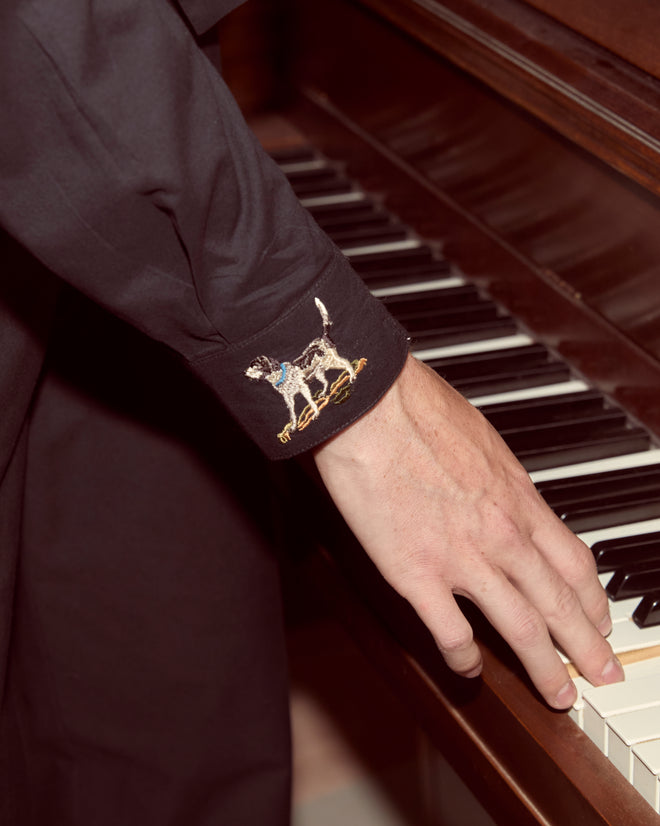 A hand plays piano keys; the Pointer Long Sleeve Shirt’s black cotton sleeve features an embroidered dog motif near the cuff.