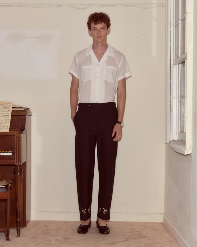 A man in a white short-sleeve shirt and Pointer Side-Buckle Trousers stands indoors beside a piano and a window.