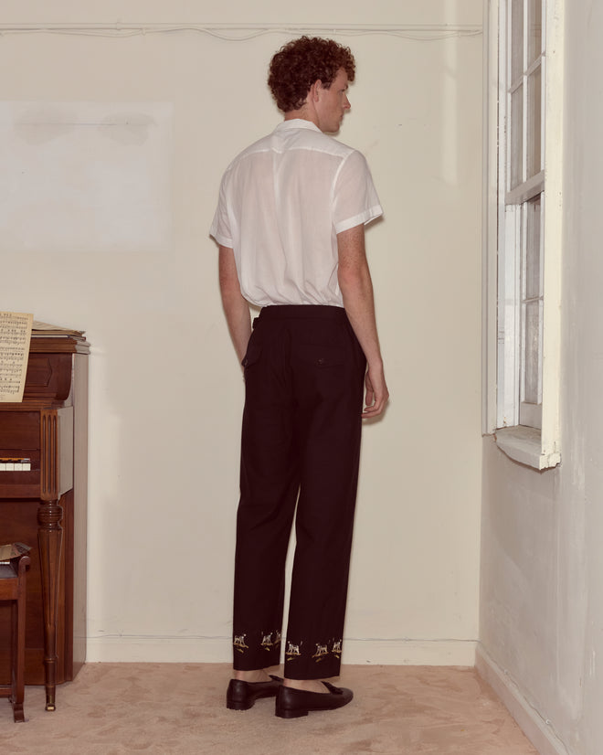 A man in a white short-sleeve shirt and Pointer Side-Buckle Trousers stands facing a corner next to a piano and a window.