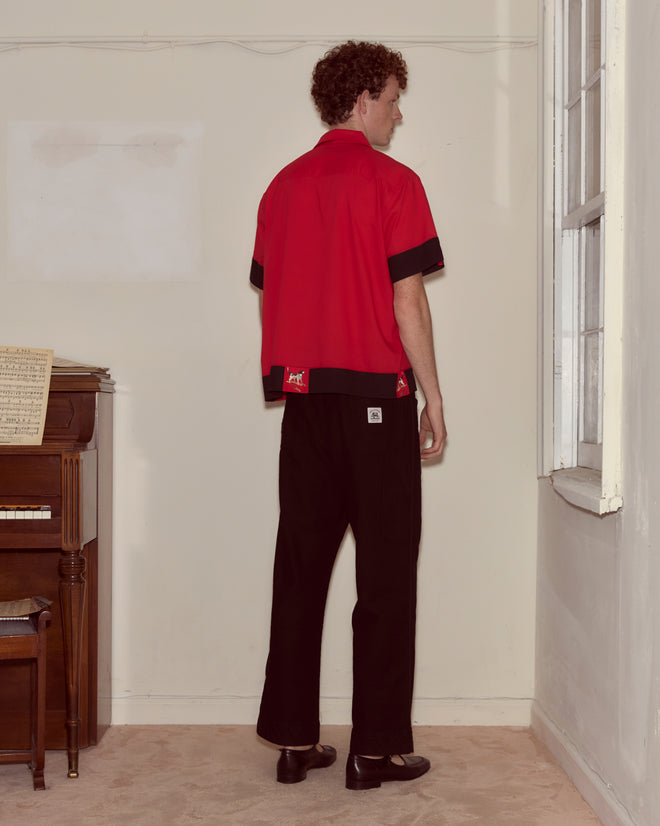 A person in a red Pointer Short Sleeve Shirt with ladderwork detailing and black pants stands facing a wall beside a piano and window.