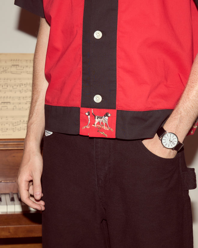 Person wearing the Pointer Short Sleeve Shirt in red and black cotton with ladderwork detailing, paired with a watch, standing by a piano with sheet music in the background.