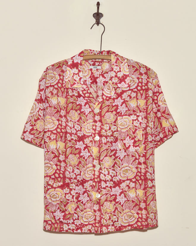 Pomegranate Leaf Short Sleeve Shirt (M/L) featuring a yellow and white floral pattern on red fabric, short sleeves, and button-up front, displayed hanging on a hook against a wall.
