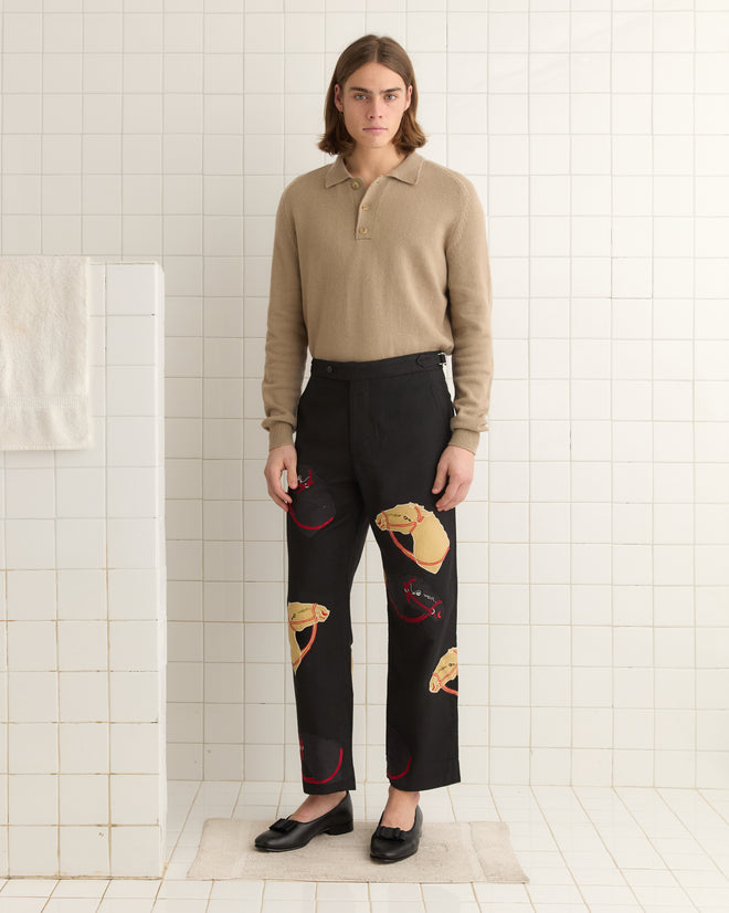 A person with long hair stands in a tiled room wearing a beige wool sweater and black Pony Appliqué Trousers adorned with taco designs.
