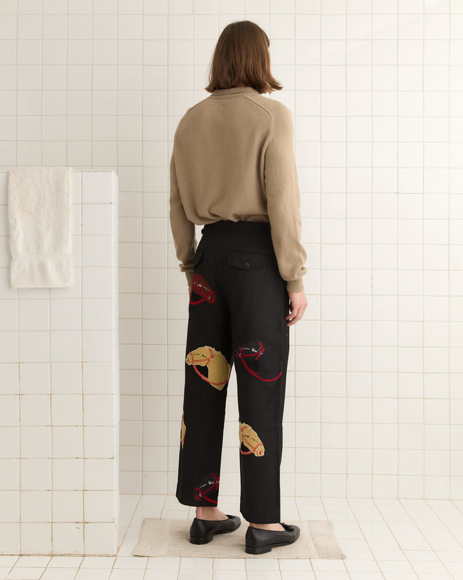In a tiled room, facing away, a person wears black Pony Appliqué Trousers paired with a beige sweater adorned with a horsehead design.
