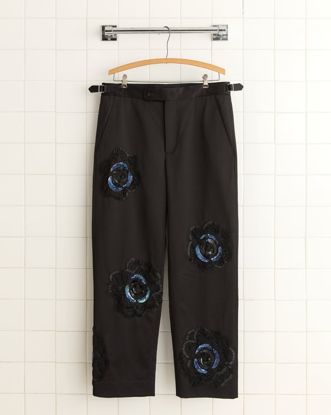 The Poppy Suiting Trousers in black, featuring blue floral patches, hang on a hanger against a tiled wall, capturing the elegance of 1940s-inspired fashion.