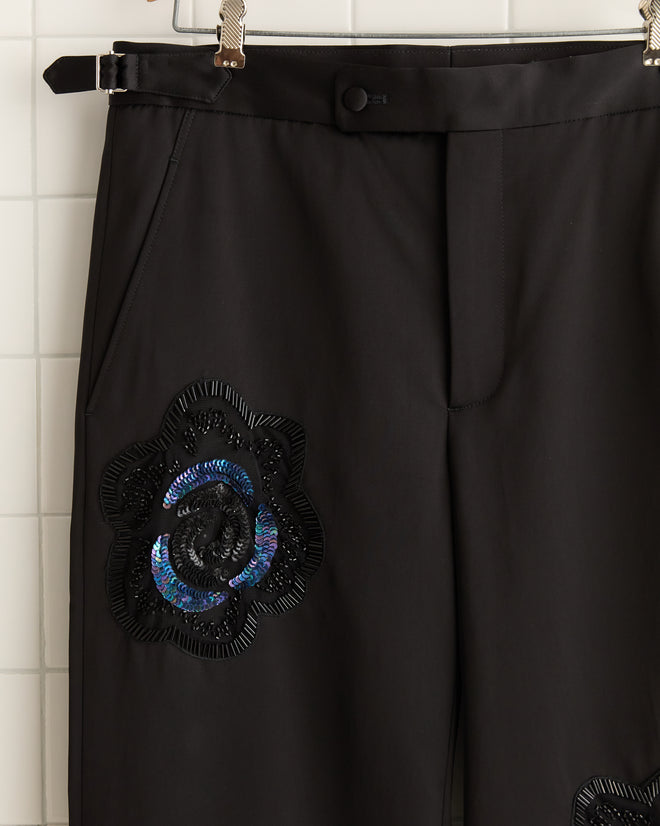 Against a tiled wall, the Poppy Suiting Trousers in black feature a blue and black floral appliqué. Made with a satin weave and inspired by 1940s trousers, they add vintage charm to any wardrobe.
