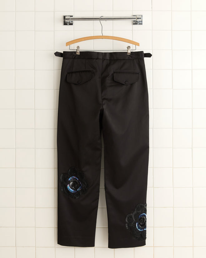 The Poppy Suiting Trousers in black, adorned with embroidered floral patches on the legs and elegantly displayed on a hanger against a tiled wall, capture a 1940s-inspired charm.