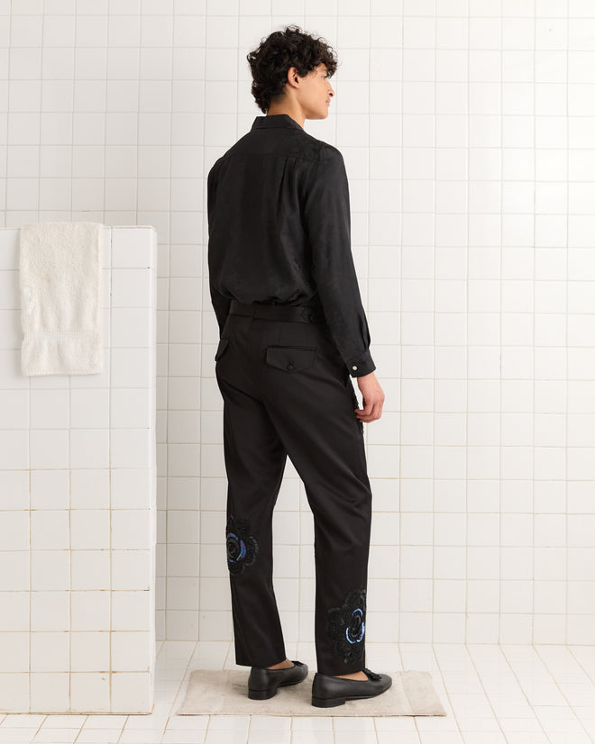 Wearing Poppy Suiting Trousers - Black, a person stands on a bath mat in a white-tiled bathroom, facing away.