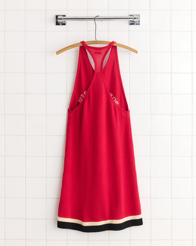 A sleeveless red Port Dress with black and white trim, inspired by early 1900s rowing singlets, hangs on a wooden hanger against a white tiled wall.
