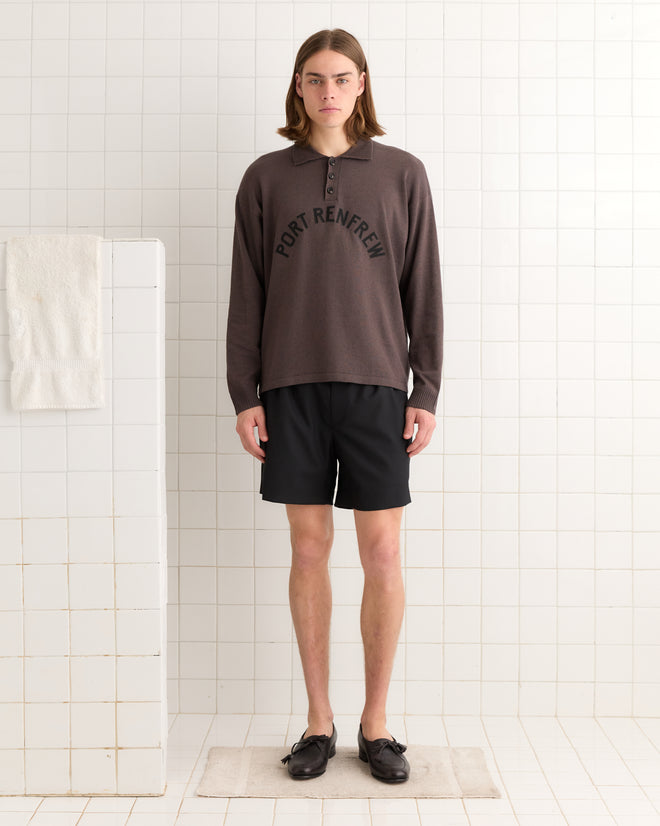 A person with long hair wearing a brown Port Polo and black shorts stands in a tiled room, where a towel hangs on a wall hook.