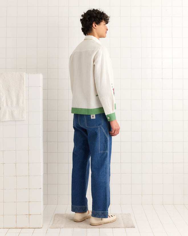 A person stands sideways in a tiled room, wearing the Primary Sailboat Long Sleeve Shirt in Green Cream, blue jeans, and white sneakers.
