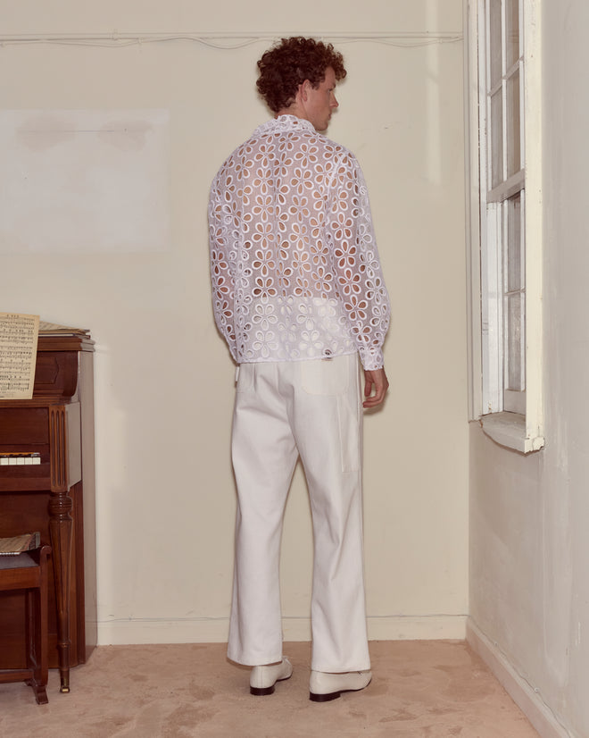 A person stands indoors, facing away from the camera, wearing white pants and the Primrose Lace Long Sleeve Shirt, featuring a sheer fabric with a delicate floral pattern.