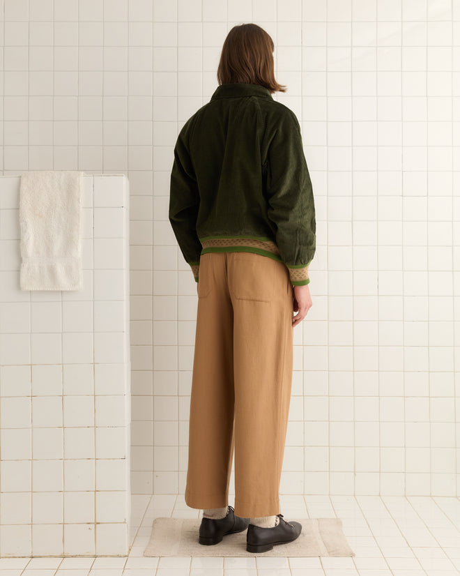 In a tiled room, someone stands facing away, wearing the Peerie Rib Banbury Jacket in forest green corduroy paired with beige pants.