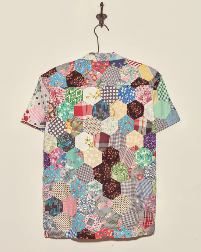 The Rainbow Fish Quilt Shirt - XS/S, a short-sleeved vintage top with a colorful patchwork hexagon design, hangs on a hook against a white wall.