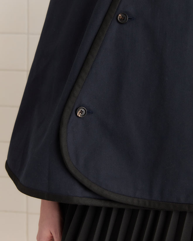 A close-up of a Waxed Herringbone Rain Cape in navy, featuring black buttons and trim, crafted from water-resistant waxed cotton, over a pleated skirt. A white tiled wall is in the background.