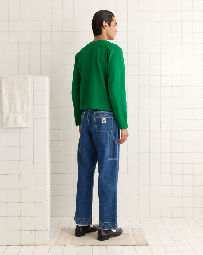A person in a green Rec Pullover and blue jeans stands on a mat in a white tiled room, facing away.