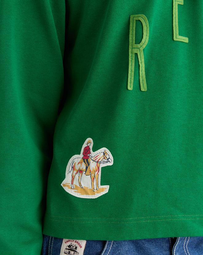 The Rec Pullover - Green is a retro-inspired shirt featuring yellow letters and a unique patch of a person in pink riding a white horse, reminiscent of vintage jerseys.