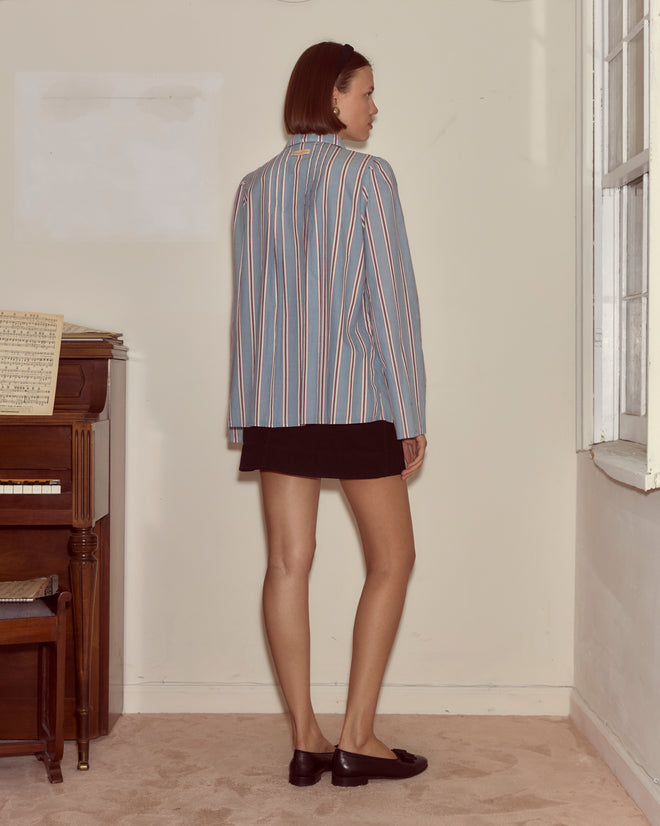 A woman wearing the Regatta Stripe Jonas Shirt and a black skirt stands by a piano in a room with beige carpet and a window.