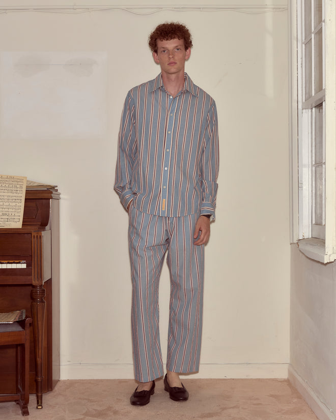 A young man in Regatta Stripe Pajama Pants, made of 100% cotton with blue stripes, stands by a piano in a softly lit, neutral-colored room.