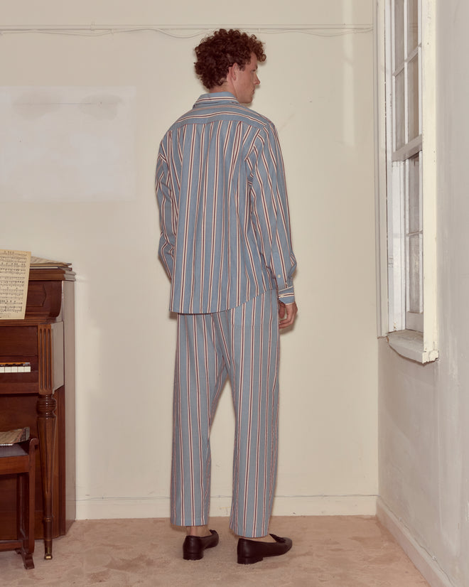 A man in Regatta Stripe Pajama Pants stands facing a wall near a piano and window in a softly lit room, his 100% cotton sleepwear adding to the cozy atmosphere.