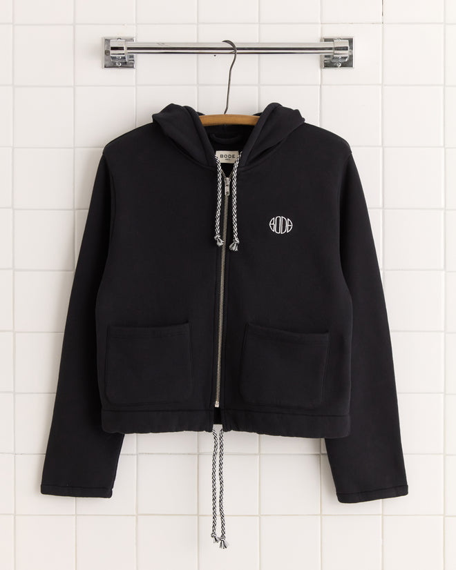 Regionals Hoodie - Black: A black cropped zip-up hoodie with front pockets, white drawstrings, and a small white logo, made from soft cotton sweatshirt material and displayed on a hanger.