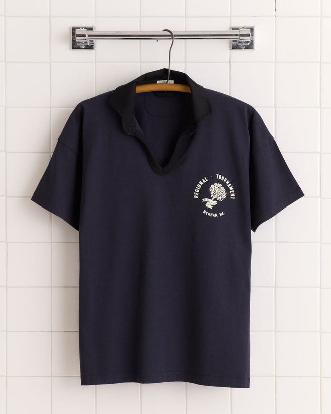 Regionals Polo: Navy blue 100% cotton polo T-shirt with black collar, white logo, and text, displayed on a hanger against a tiled wall.
