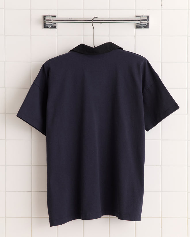 The Regionals Polo, crafted from 100% cotton in navy blue, hangs on a hanger with its back facing out against a white tiled wall.
