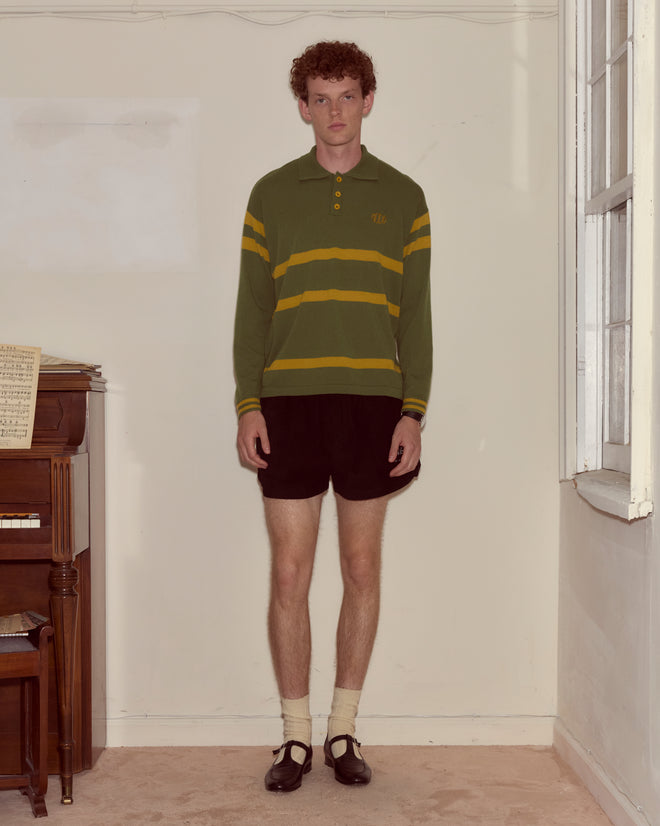A young man wearing the Regis Polo in green virgin wool and black shorts stands indoors by a piano and a window.