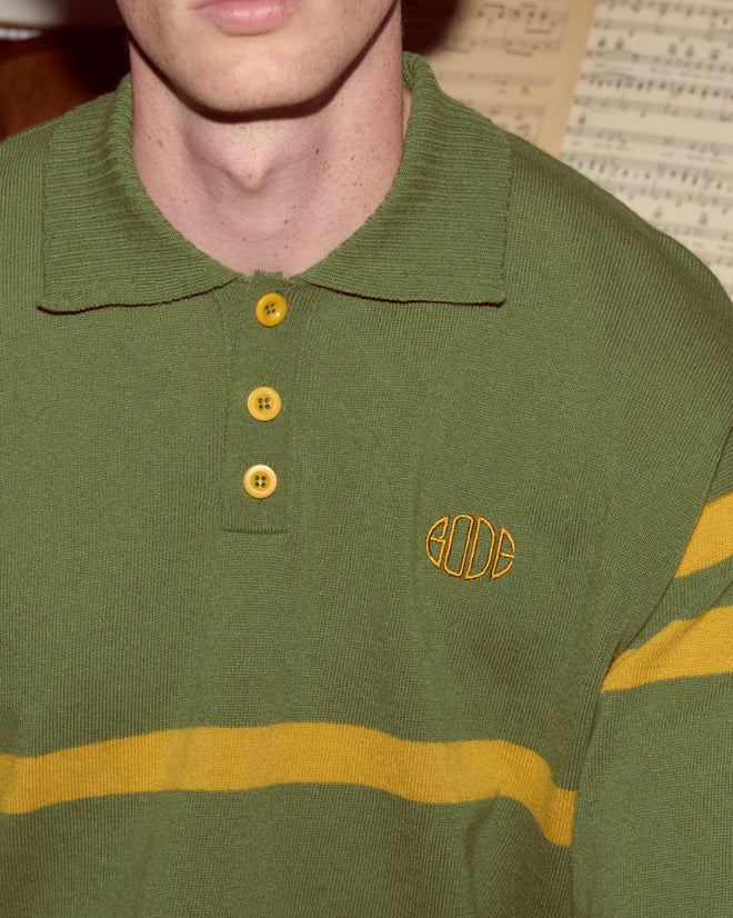 A person wears the Regis Polo, a green virgin wool sweater with yellow stripes and Bode monogram, with a music sheet in the background.