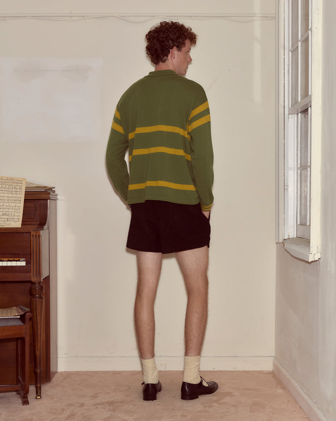 A person in a green striped Regis Polo sweater and shorts stands facing a corner near a piano and window.