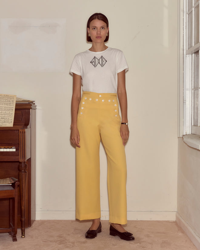 Woman in a white t-shirt and yellow Riding Twill Sailor Trousers stands by a piano in a cream-walled room with a large window.