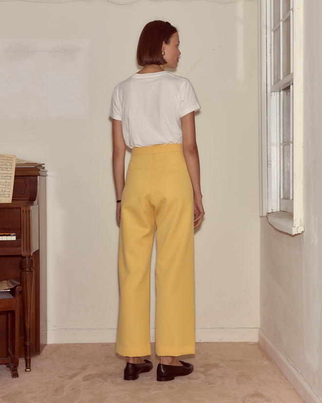 Woman stands indoors facing away, wearing a white shirt and Riding Twill Sailor Trousers in yellow, near a piano and window.