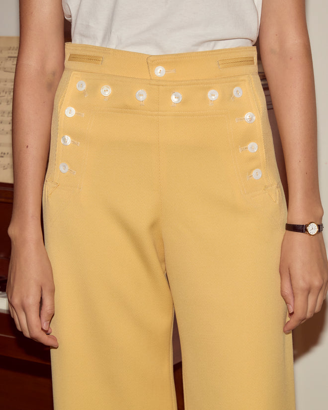A person wearing Riding Twill Sailor Trousers in yellow and a crisp white shirt, standing indoors.