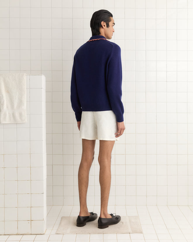 A person wearing the Rookie Polo, featuring a purple color with candy-striped trim, pairs it with white shorts and black shoes while standing in a tiled room facing away.