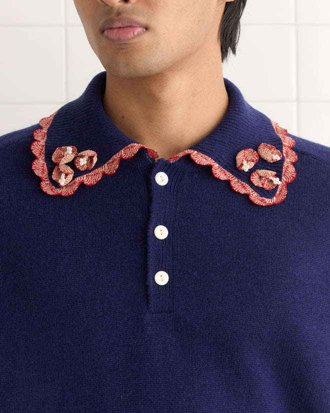 A person wearing the Rookie Polo, a blue sweater with a red scalloped collar and white buttons, showcases delicate pearl seed beads along the edges.