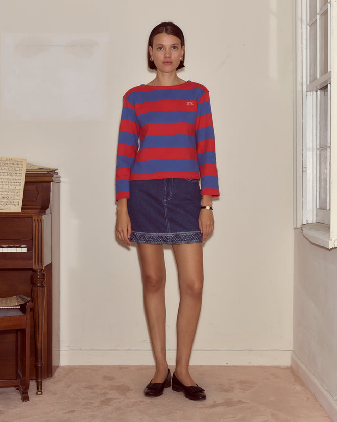 A woman wears the Clubhouse Long Sleeve, a red and blue striped 100% cotton tee, paired with a denim skirt while standing indoors by a piano and window.