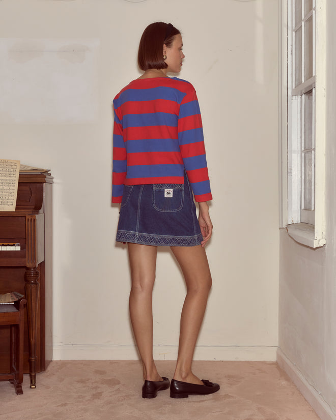 A woman stands by a window, facing away from the camera, wearing the Clubhouse Long Sleeve in red and blue stripes paired with a denim skirt.