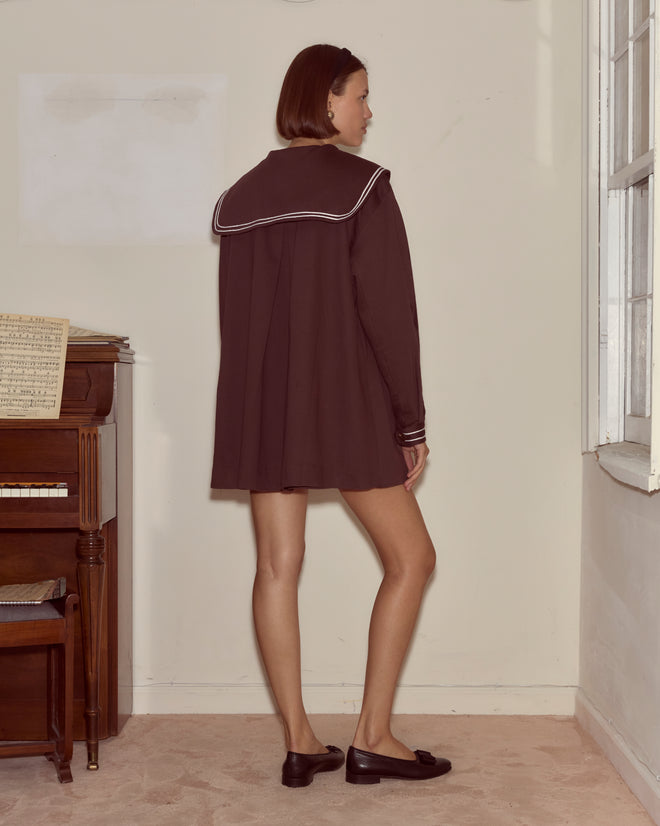 A person wearing the Sailor Coat - Brown, inspired by a 1960s reproduction, stands indoors near a piano with their back to the camera.