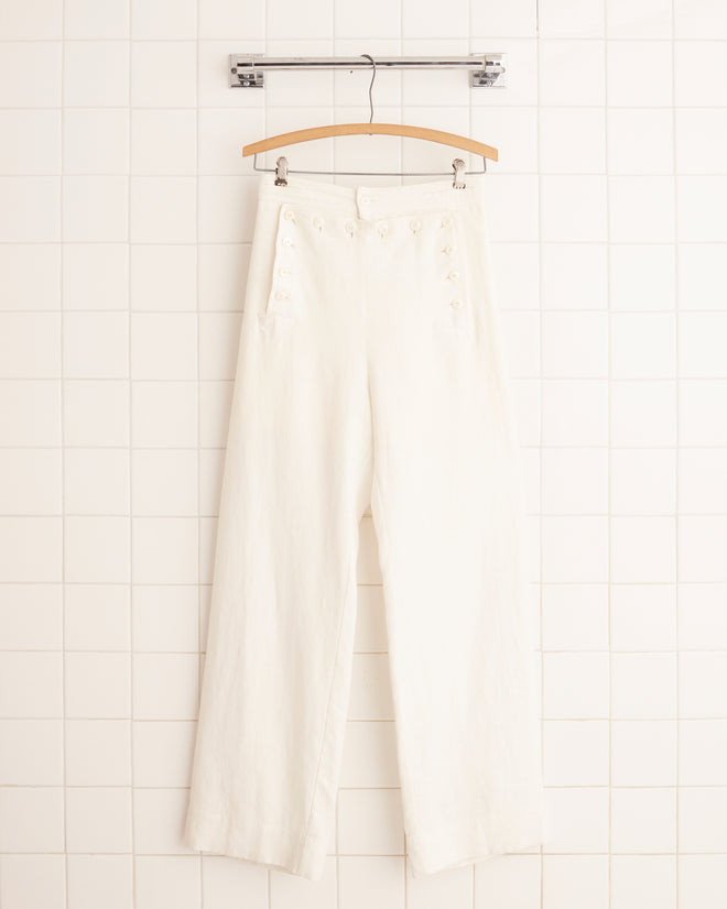 The Linen Sailor Trousers in white, hanging gracefully against a tiled wall, evoke timeless elegance and vintage charm from the 1940s.