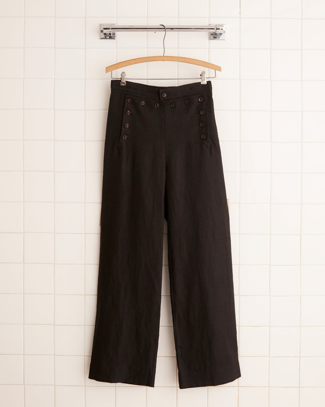 Linen Sailor Trousers - Black, featuring button details and wide legs, hang on a wooden hanger against a tiled wall.