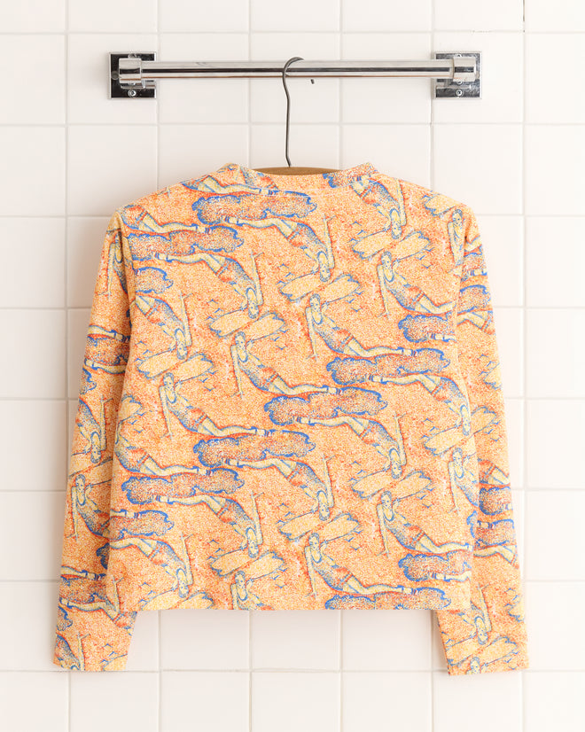 The Sandbank Long Sleeve Tee, with its vibrant orange and blue pattern, hangs against a white tiled wall, echoing the lively hues of a 1960s-1970s design.