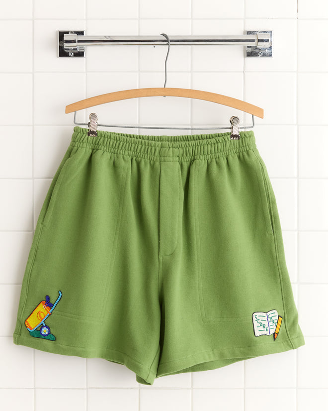 Score Card Shorts: green sweatshorts with embroidered juice box and notepad with pencil, inspired by Canadian rugby shorts, displayed on a wooden hanger.