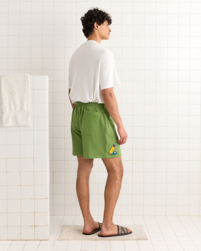 Man in a white shirt and green Score Card Shorts stands on a bath mat in a tiled bathroom, facing sideways.