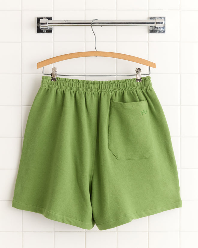 Score Card Shorts in green, featuring an elastic waistband and back pocket, displayed on a wooden hanger against white tiles.