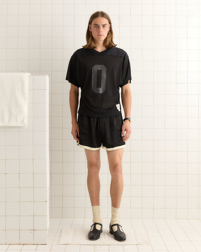 A person wearing the Scrimmage Jersey - Black stands in a tiled room, with a towel hanging on a partition to the left.