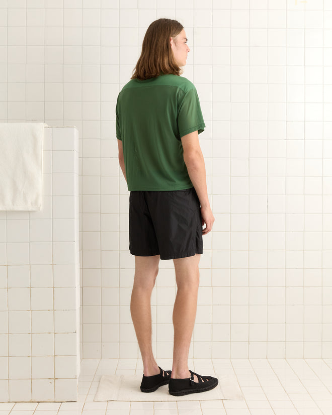A person with long hair, seen from behind, wears the Scrimmage Jersey - Green and black shorts in a white tiled room.